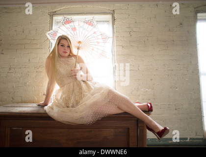 A 16-year-old Caucasian teenager dressed in a white, 1960's vintage lace dress sits on an antique piano holding an umbrella. Stock Photo