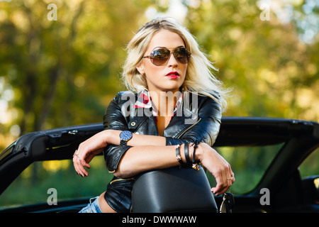 Blond woman in leather jacket and jeans shorts in convertible car