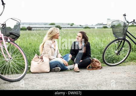 Young female friends with bicycles conversing while sitting on country road Stock Photo