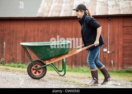 Side view of female farmer pushing wheelbarrow on rural road Stock Photo