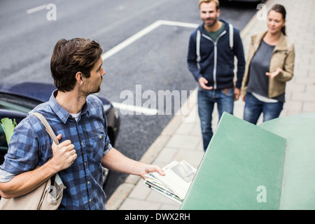 Man putting newspaper in recycling bin while looking at friends on sidewalk Stock Photo