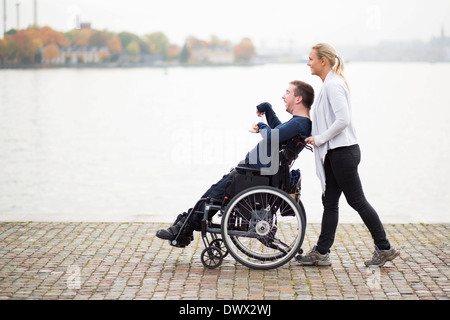 Caretaker pushing disabled man on wheelchair along lake Stock Photo