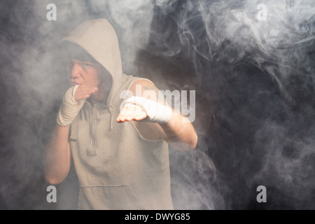 Young boxer working out in a smoky room in a hooded grey top standing with his bandaged hands raised waiting and watching Stock Photo