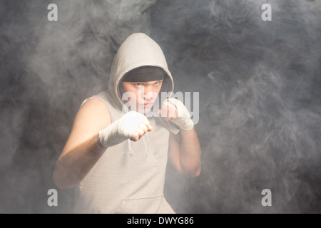 Young boxer fighting in a dark smoke filled atmosphere standing with his fists raised at the ready and a grim intense expression of determination, with copyspace. Stock Photo