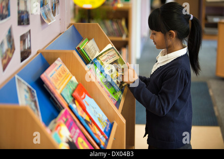 An Asian primary school pupil reading next to a book shelf in the UK Stock Photo