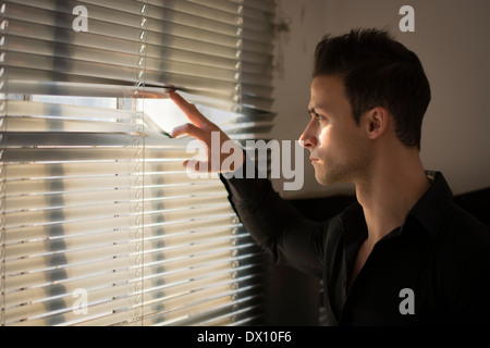 Profile of young man peeking through venetian blinds in a dark room Stock Photo