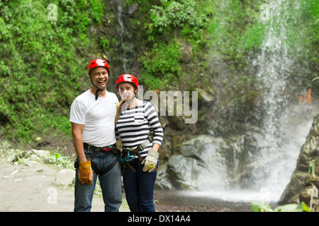 A man and a woman stand side by side in front of a cascading waterfall, both wearing red helmets, climbing harnesses, and gloves. The man is in a white t-shirt and jeans, smiling broadly, while the Stock Photo