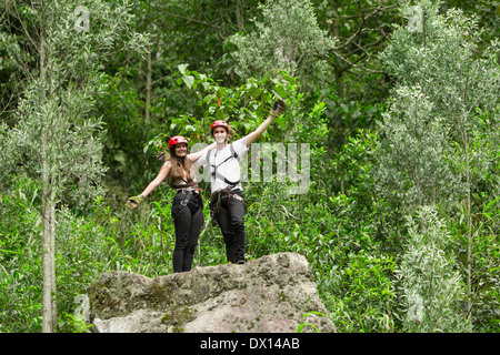 A man and a woman wearing red helmets, climbing harnesses, and gloves stand together on a large moss-covered rock in a lush green forest. Both are smiling and raising their arms in a celebratory Stock Photo