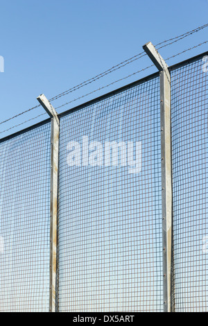 Tall wire mesh security fence on concrete posts, topped with three strands of barbed wire at a 45 degree outward-facing angle Stock Photo