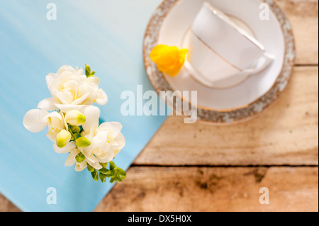 White freesias on blue mat on rustic wooden table with blurred vintage teacup and yellow freesia in background Stock Photo