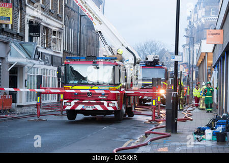 Firefighter in yellow helmet stands on red engine at base of telescopic ladder by fire damaged building, crew & trucks behind - Harrogate, England, UK Stock Photo