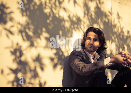 A fashionable young man sitting by a wall in the shade on a sunny day Stock Photo