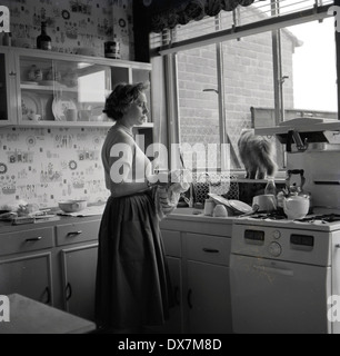 Historical picture from the 1950s showing a housewife standing by her kitchen sink holding a tea towel drying the dishes, with a cat on the window sill, England, UK. Stock Photo