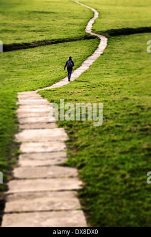 Woman walking on a long flagstone pathway snaking through a grassy field in the Yorkshire Dales, England, UK. Stock Photo