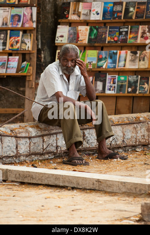 South Southern India Kerala Fort Cochin Kochi quayside grey gray haired old man male fisherman sitting sits on wall bookstall paperbacks novels sunny Stock Photo