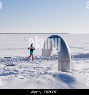Young Woman Running in the Snow. A woman runs by on a snowy path, out on the ice a person views the scene, windmills in distance Stock Photo