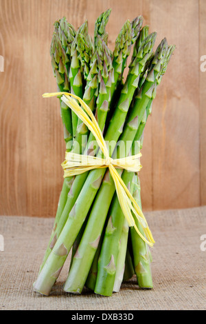 Bunch of fresh green asparagus spears tied with yellow raffia stand on a rustic hessian cloth by vintage wood background, UK Stock Photo