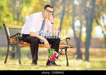 Sad young man holding a bouquet of flowers and sitting on a bench in a park Stock Photo
