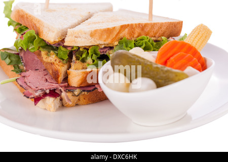 Delicious thinly sliced beef pastrami club sandwich with fresh curly lettuce served with a small dish of pickled vegetables on a white plate, isolated on white Stock Photo