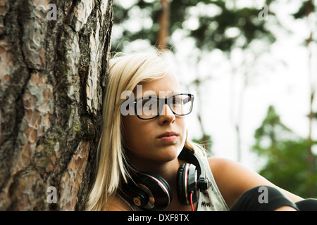 Close-up portrait of girl wearing eyeglasses, sitting next to tree in park, with headphones around neck, Germany Stock Photo
