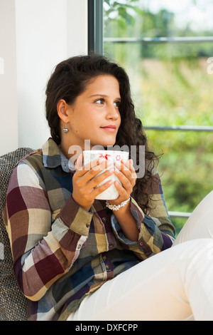 Teenage girl sitting next to window, holding cup, Germany Stock Photo