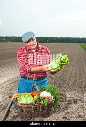 Farmer kneeling in field with basket of fresh vegetables, smiling and looking at lettuce, Hesse, Germany