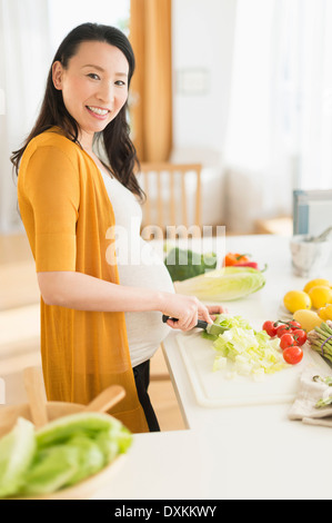 Portrait of pregnant Japanese woman slicing vegetables in kitchen Stock Photo