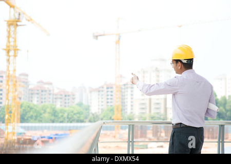 Portrait of a smiling Asian Indian male contractor engineer with hard hat pointing to construction site. Stock Photo