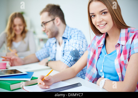 Smart girl looking at camera while carrying out written task with her groupmates on background Stock Photo