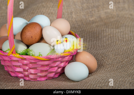Pink Easter basket filled with colorful eggs and two decorative chickens, closeup. Burlap background. Stock Photo