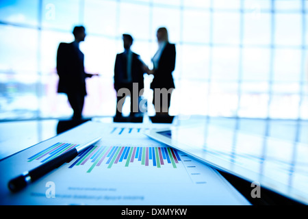 Digital tablet and financial document with pen at workplace on background of three business partners interacting Stock Photo
