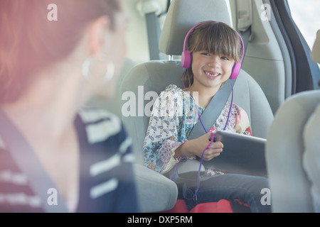 Mother turning and smiling at daughter with headphones and digital tablet in back seat of car Stock Photo