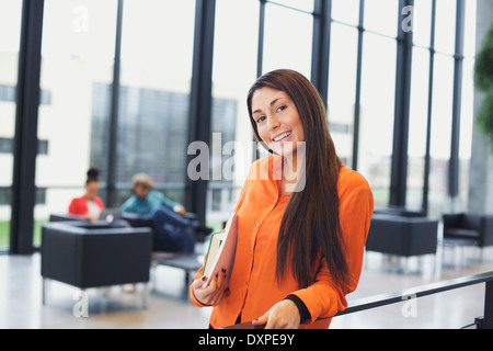 Pretty young student holding books standing by a railing looking at camera smiling. Caucasian female in university lobby. Stock Photo