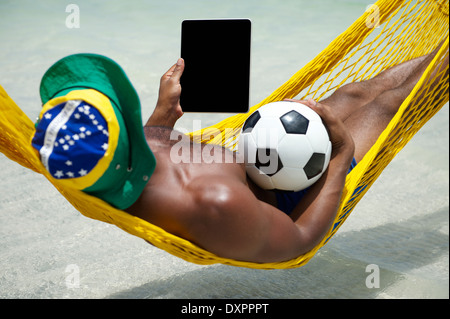 Brazilian man using tablet computer with football soccer ball relaxing in hammock on beach over the sea Stock Photo