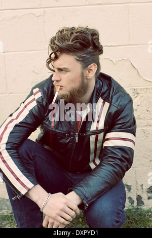 A bearded man in pompadour vintage hairstyle smoking a cigarette squatting down, an edgy vintage concept Stock Photo