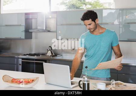 Man with newspaper using laptop in kitchen Stock Photo