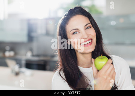 Portrait of smiling woman eating apple in kitchen Stock Photo
