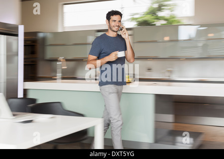 Smiling man drinking coffee and talking on cell phone in kitchen Stock Photo