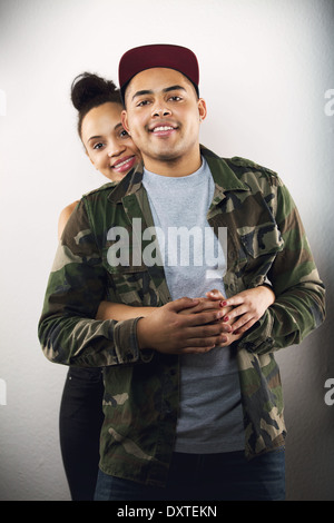 Young lady hugging her boyfriend from behind. Lovely young couple in love together on gray background. Stock Photo