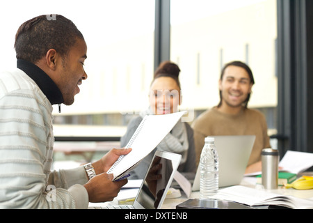 Young man reading an assignment with classmates laughing in background. Young students studying together in library. Stock Photo