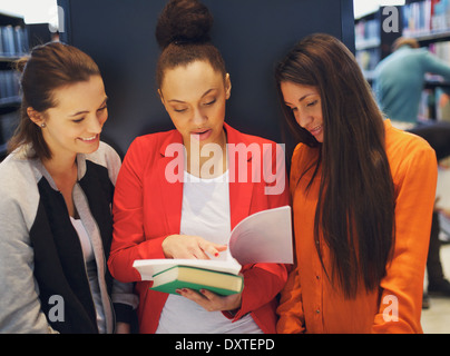 Three young female students standing in the library and looking at a book together. university students reading reference books. Stock Photo