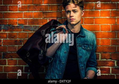 Young Caucasian male model in denim jacket posing against brick wall Stock Photo