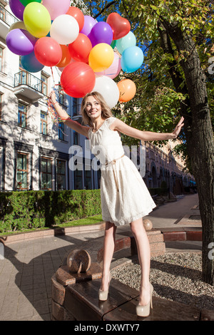 pretty woman with long legs in white summer dress holding a bunch of multicolored balloons stands on bench in city park Stock Photo