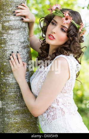 Young woman with flower wreath as headdress poses at a tree trunk