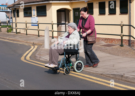 An elderly woman in a wheelchair with her carer or assistant crossing road. Public disabled toilets in background. Stock Photo