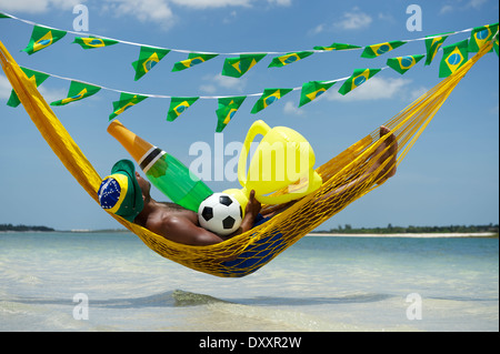 Brazilian soccer player celebrating by relaxing in beach hammock with his football, champagne bottle, and trophy Stock Photo