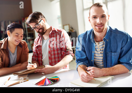 Smiling guy with pencil and notepad looking at camera with his friends discussing palette on background Stock Photo