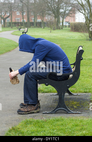 Male wearing hoodie sitting on park bench drinking alcohol Stock Photo