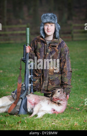 A young male deer stalker with a dead deer and a rifle in a grass field at dusk Stock Photo