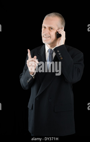 An agreeable businessman wearing a black suit smiling while speaking on mobile phone, on black background Stock Photo